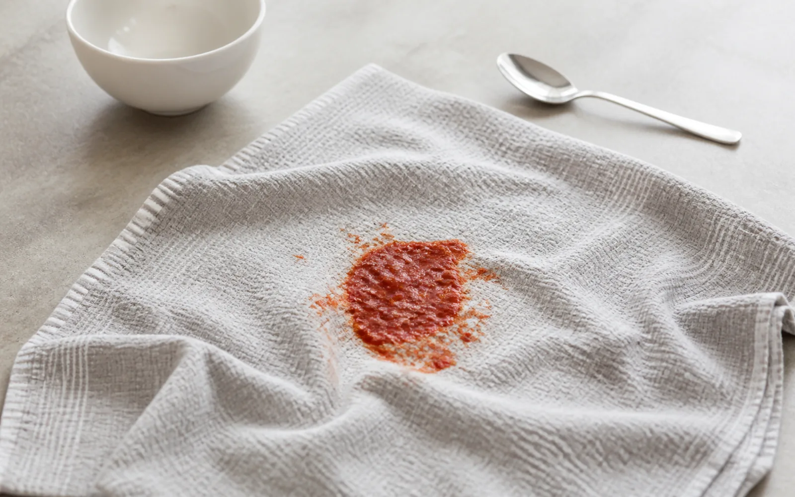 A fresh red tomato sauce spill centred on a light grey woven tea towel beside a plain white bowl and stainless steel spoon.